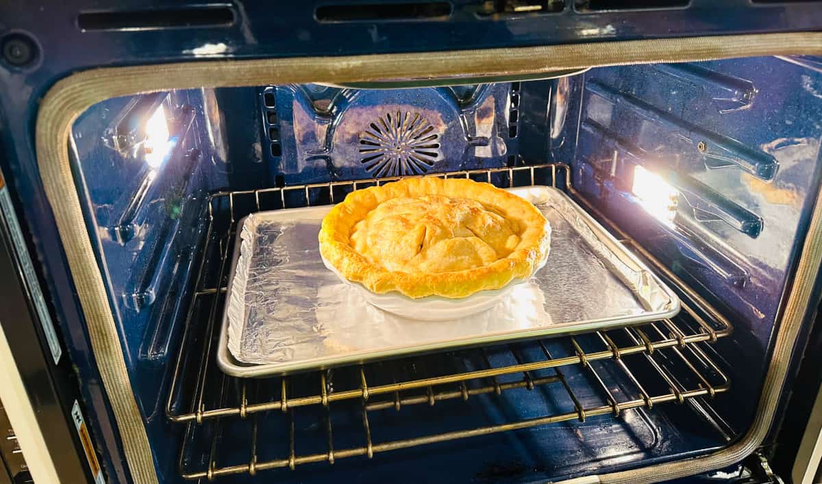Old fashioned apple pie baking on a metal cookie sheet covered in tinfoil in a blue oven.