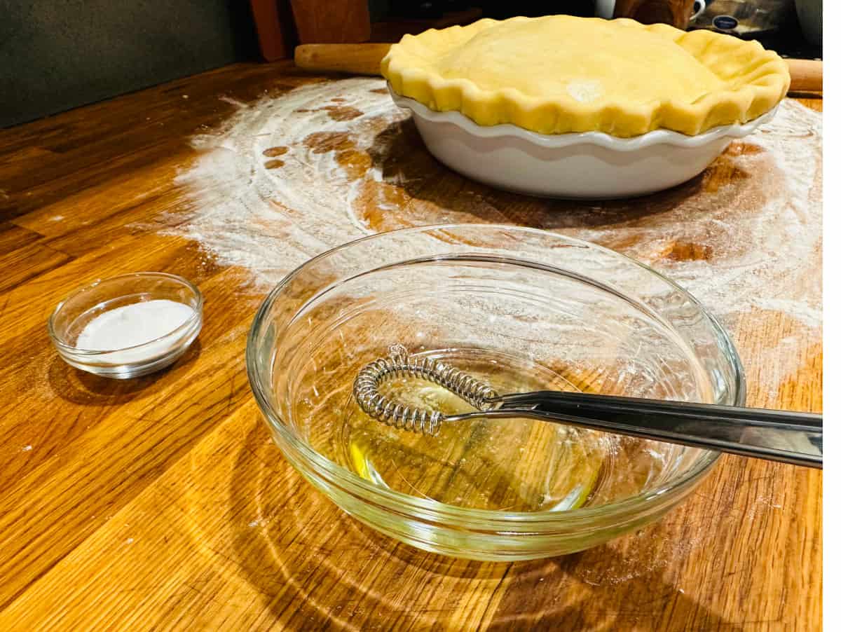 Egg white in a glass bowl with small whisk next to small glass bowl of sugar and unbaked pie in a white pie plate.