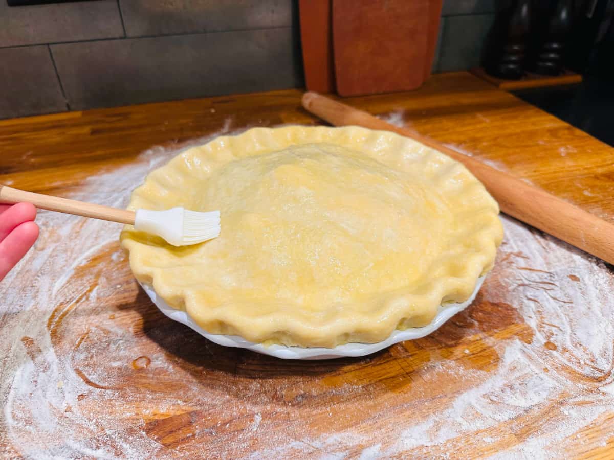 White brush applying beaten egg white on top of unbaked old fashioned apple pie.