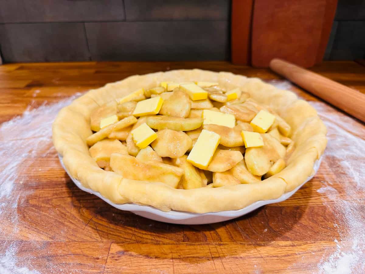 Small pieces of butter on top of sliced apples and unbaked pie dough in a white pie plate.