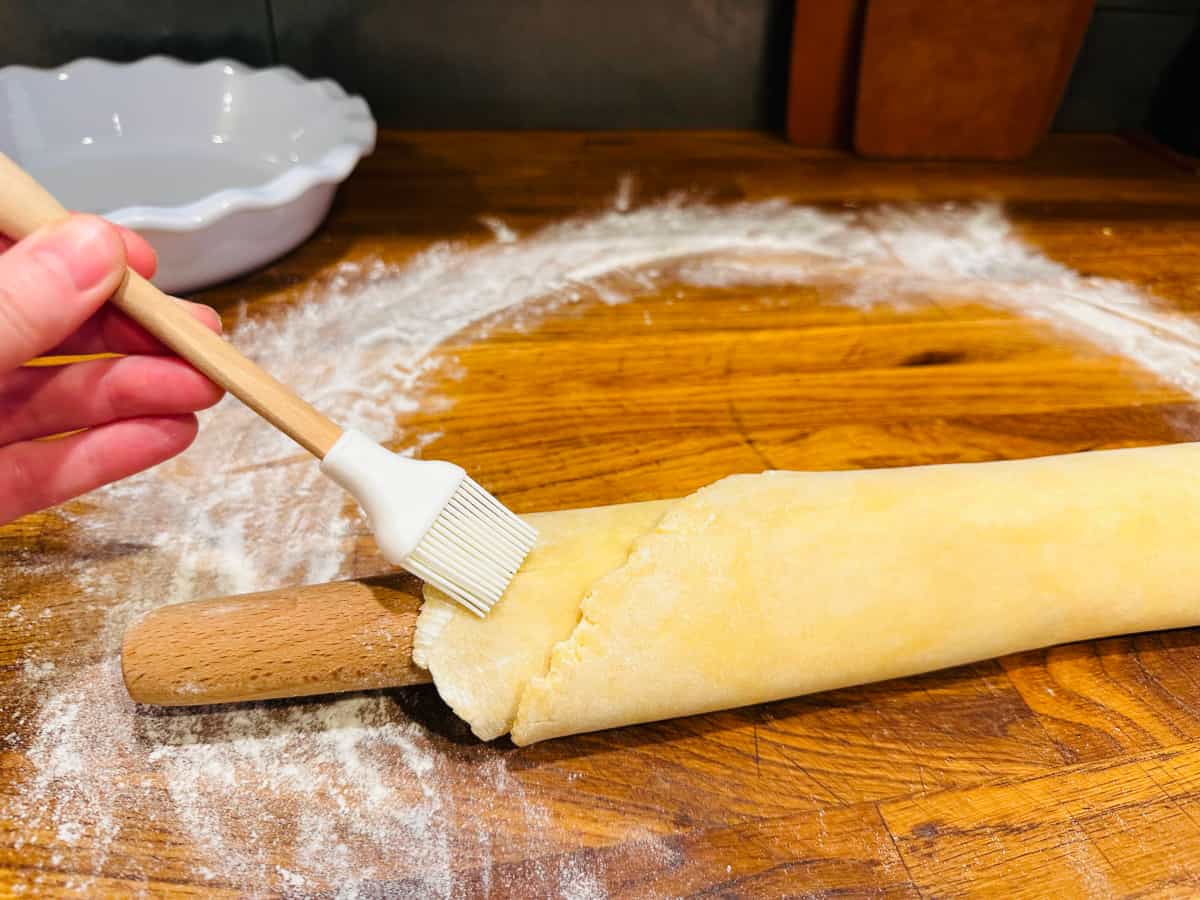 Pie dough gathered up around a rolling pin and a white brush dusting flour off the edges.