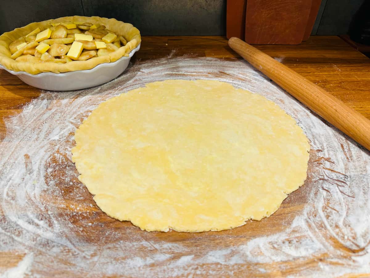 Pie dough rolled out into a big circle between pie plate full of apples and a rolling pin.