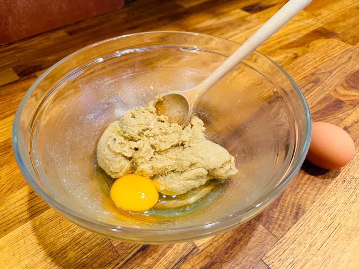 Glass bowl containing mixture of butter and sugar, one egg, and a wooden spoon.