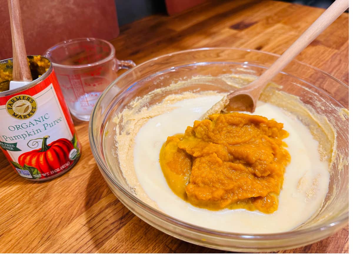 Empty can of pumpkin puree and pyrex measuring cup next to glass bowl of pumpkin and milk on top of wet ingredient mixture.