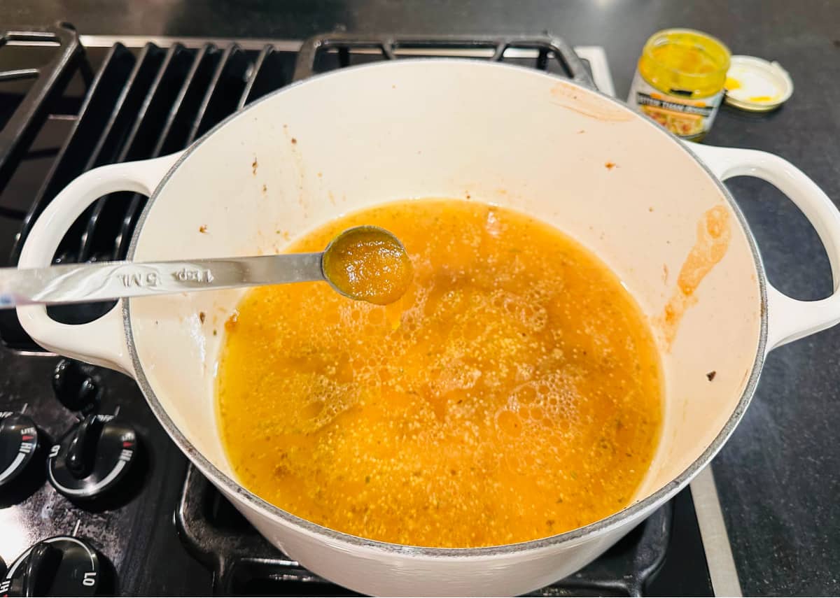 Measuring spoon full of chicken bouillon over chicken broth in a white pot.