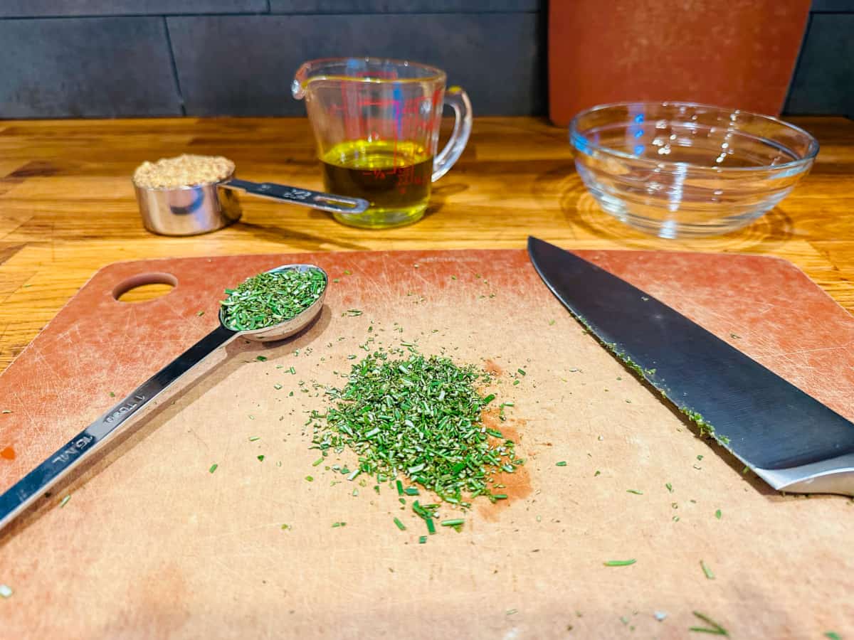 Chopped rosemary on a cutting board with a measuring spoon and butcher knife.