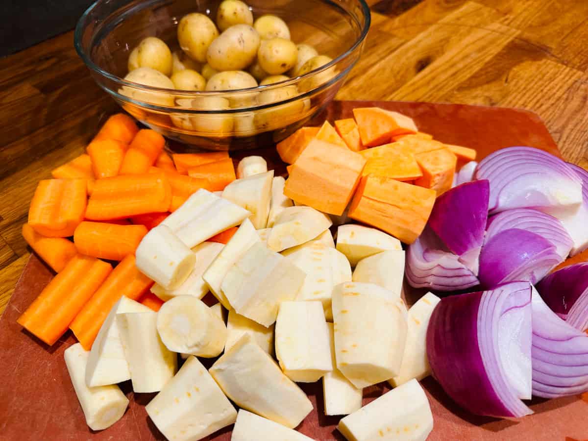 Chopped carrots, parsnips, yams, and red onions next to a glass bowl filled with small potatoes.