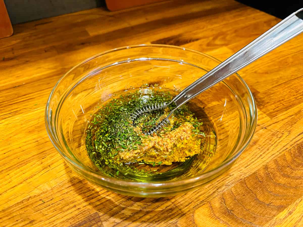 Chopped rosemary, mustard, and olive oil in a glass bowl with a small whisk.