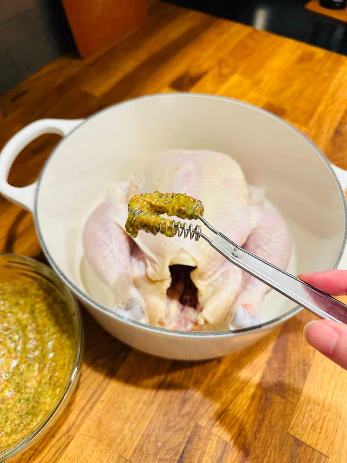 Magic whisk dipped in chicken seasoning and a whole raw chicken in a white pot.