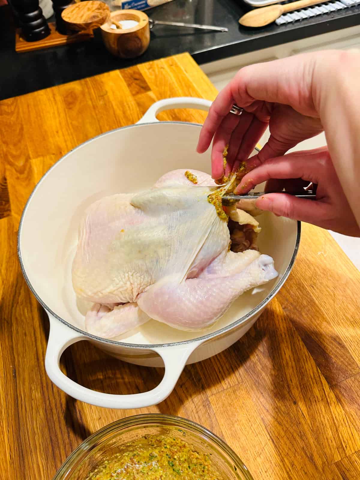 Two hands maneuvering magic whisk under the skin of a whole chicken in a white pot.