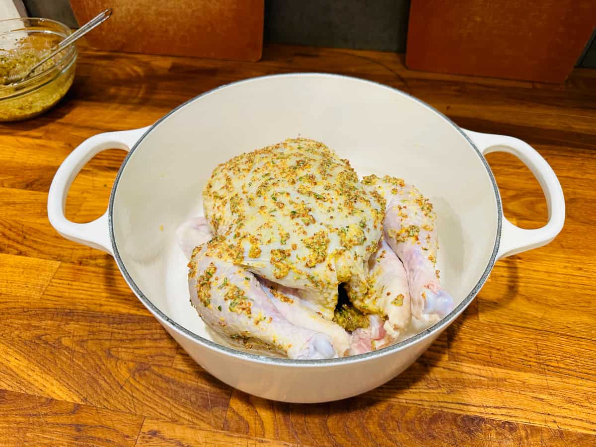 Whole raw chicken covered in chopped rosemary and mustard in a white pot.