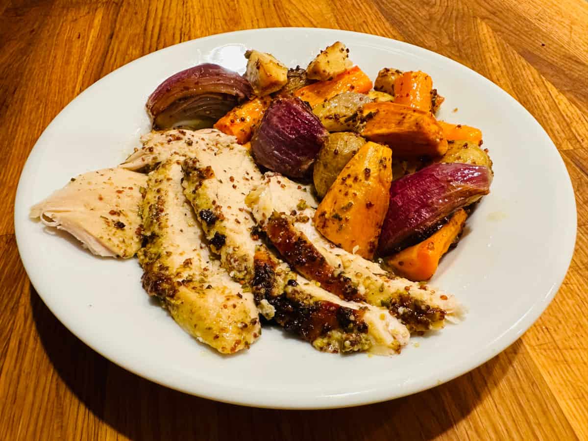 Slices of roasted chicken with roasted carrots, potatoes, yam, and red onion on a white plate.