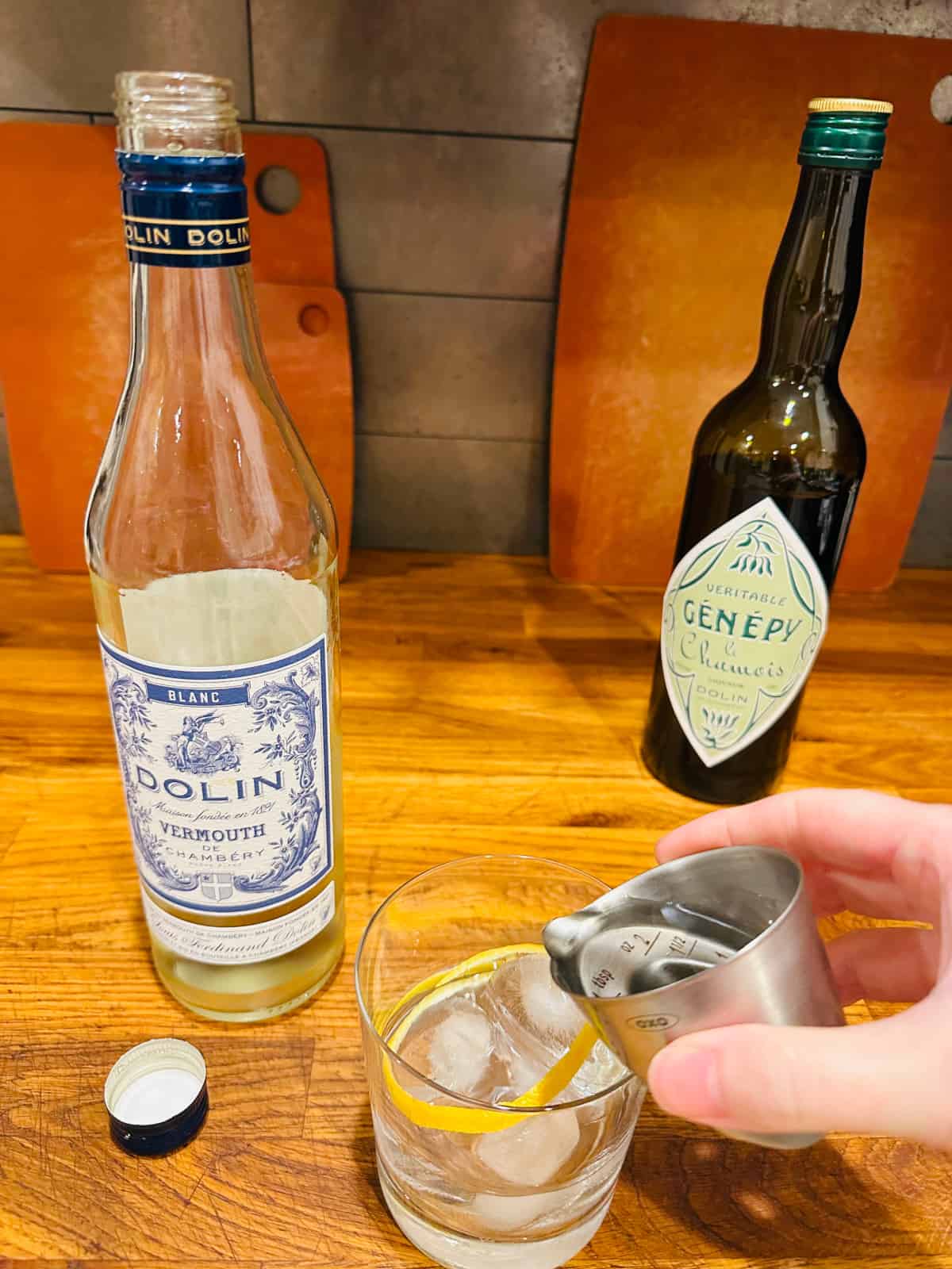 Hand pouring clear liquid from a steel measuring jigger into an old fashioned glass next to a bottle of Blanc vermouth.