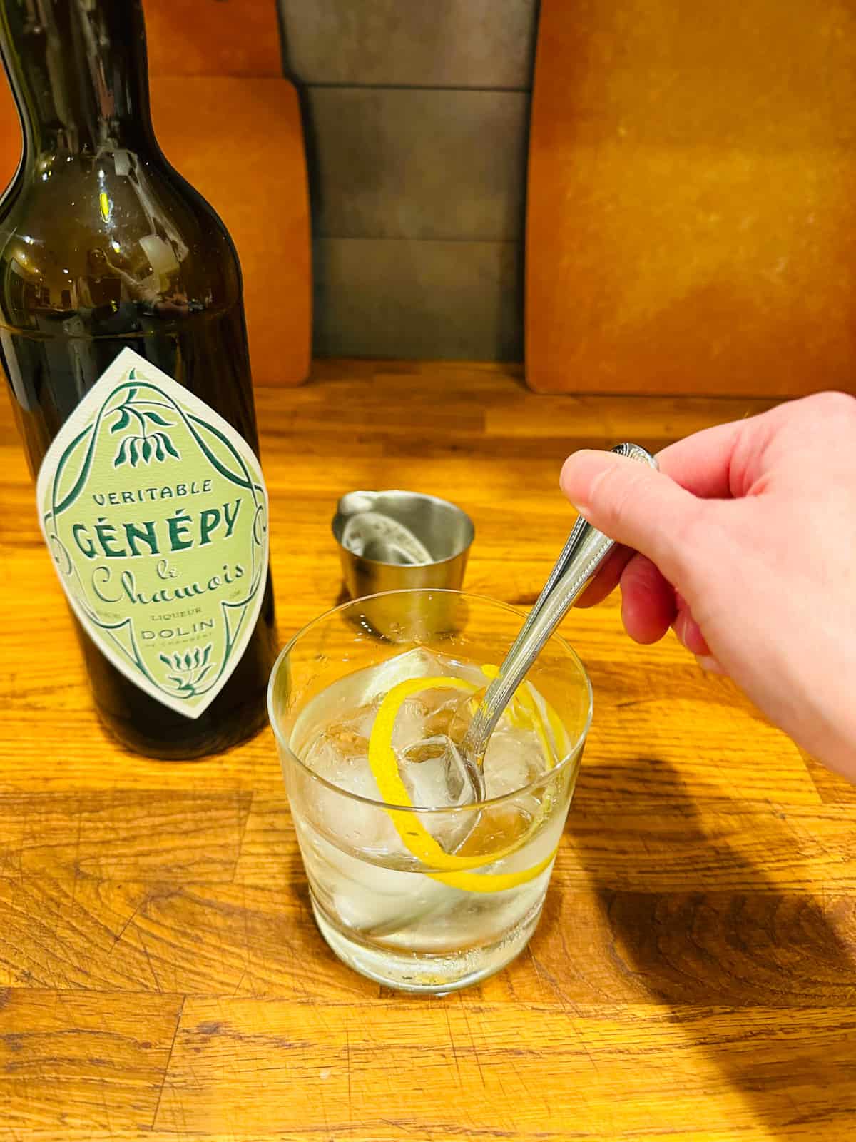 Hand stirring a white Negroni with a metal spoon next to a bottle of genepy.
