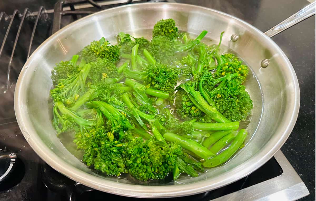 Broccolini pieces steaming in boiling water in a large skillet.
