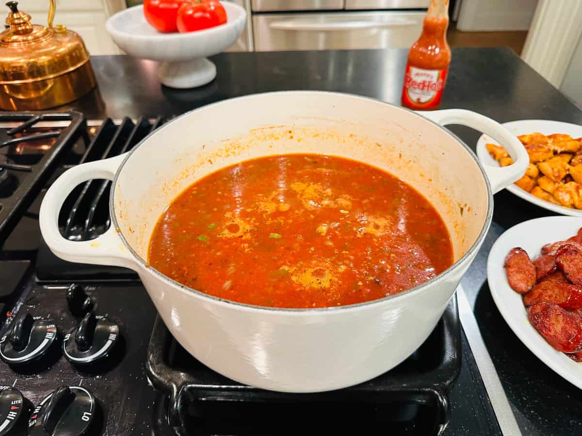 White pot filled with red liquid simmering on a stove next to a bottle of hot sauce and white plates holding browned meat.