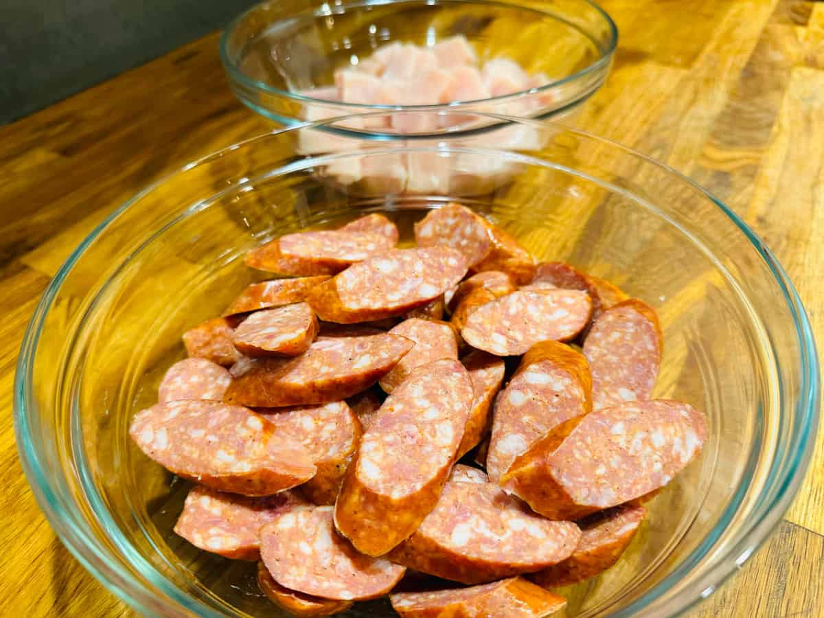 Raw sliced chorizo in a glass bowl in front of raw pieces of chicken in a glass bowl.