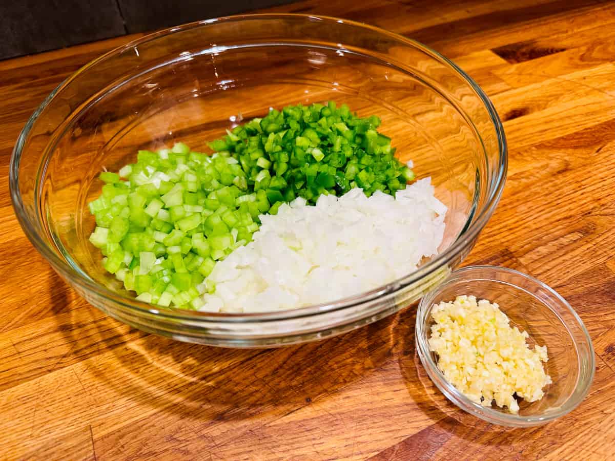 Chopped onion, celery, and green pepper in a glass bowl next to minced garlic in a small glass bowl.