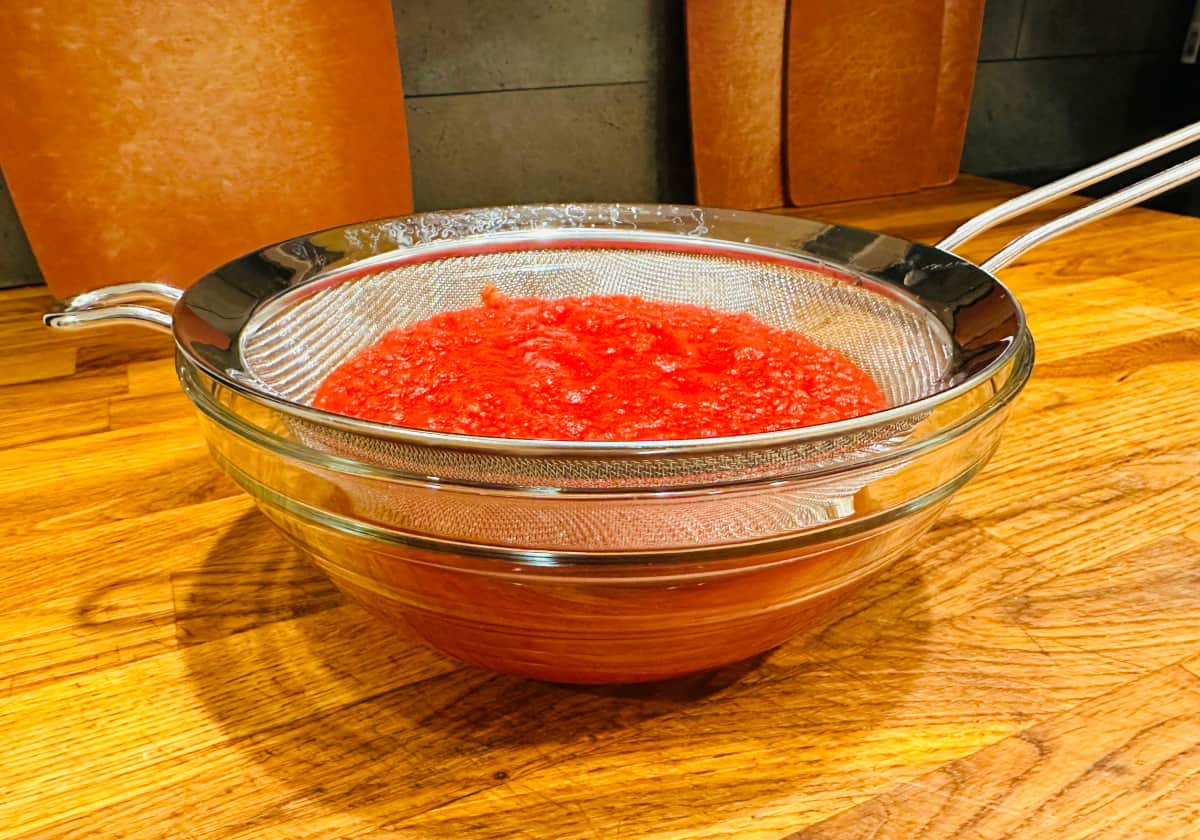 Finely chopped tomatoes in a metal strainer sitting over a glass bowl of tomato juice.