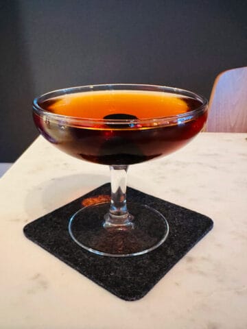 Nordic Spring (AKA Aquavit Manhattan) cocktail in a coupe glass on a dark gray coaster sitting on a white marble table.