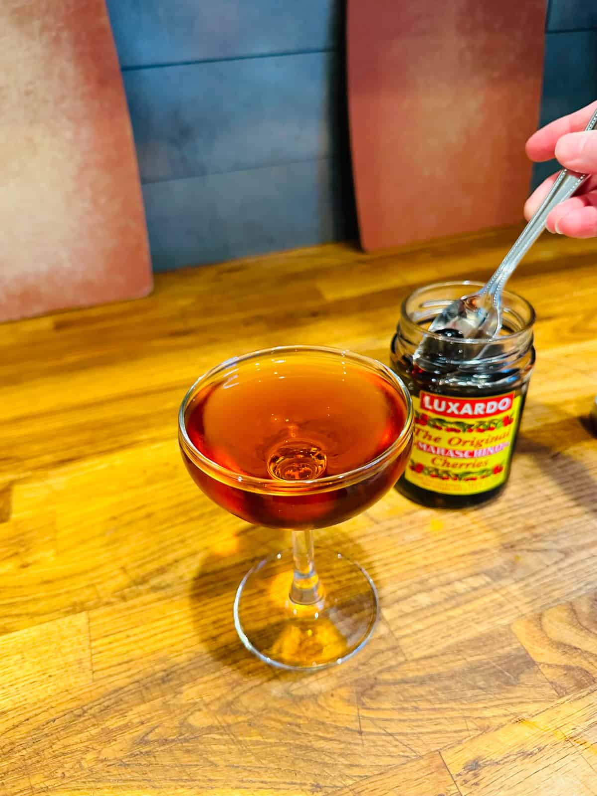 Coupe glass filled with brown cocktail next to jar of maraschino cherries.