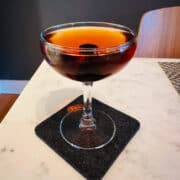 Nordic Spring (AKA Aquavit Manhattan) cocktail in a coupe glass on a dark gray coaster sitting on a white marble table.