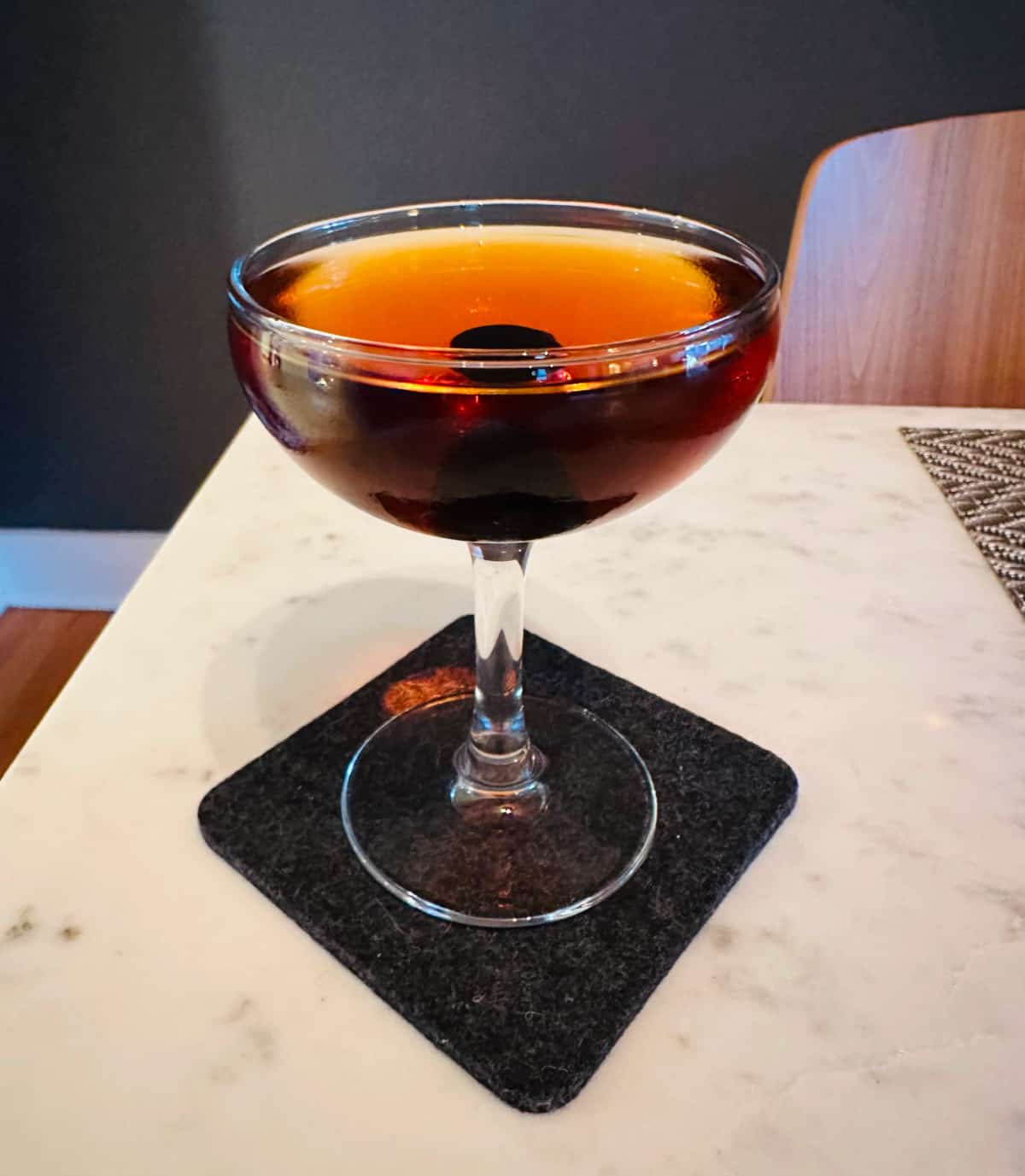 Nordic Spring (AKA Aquavit Manhattan) cocktail in a coupe glass on a dark gray coaster sitting on a white marble table.