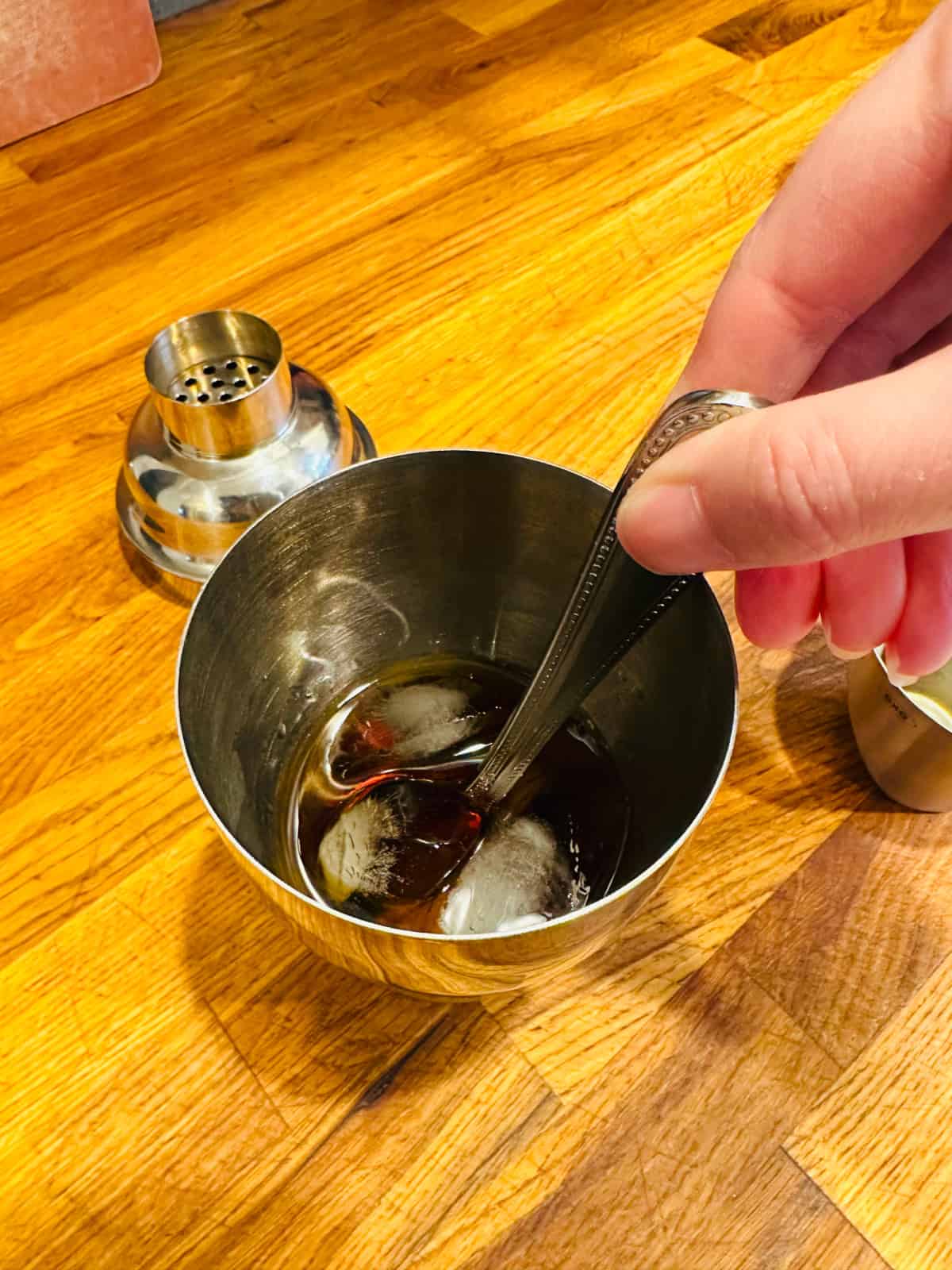 Hand stirring a spoon in a cocktail shaker filled with ice and an amber colored liquid.