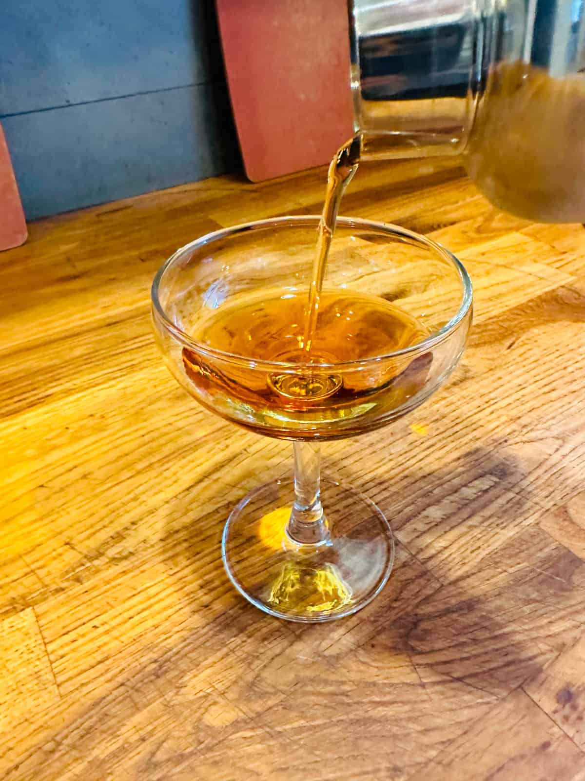 Cocktail shaker pouring amber colored liquid into a coupe cocktail glass.