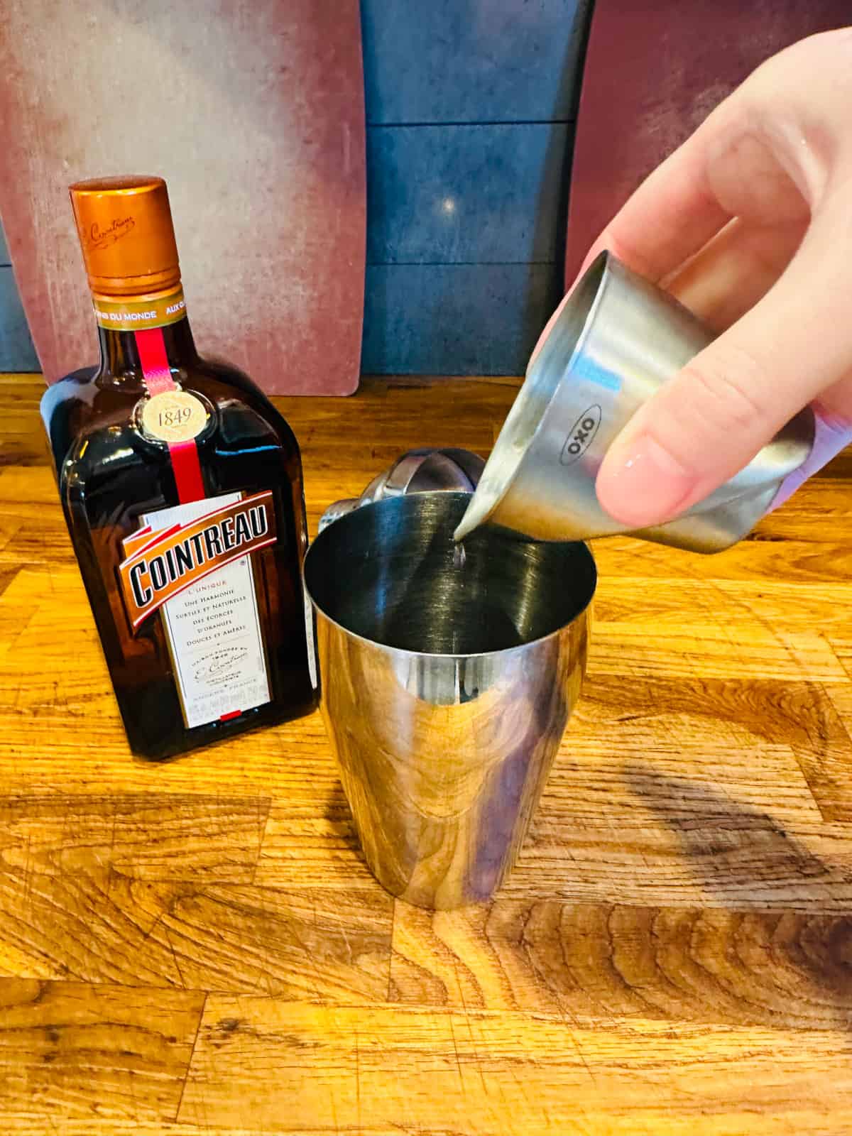 Hand pouring cointreau from metal measuring cup into cocktail shaker.