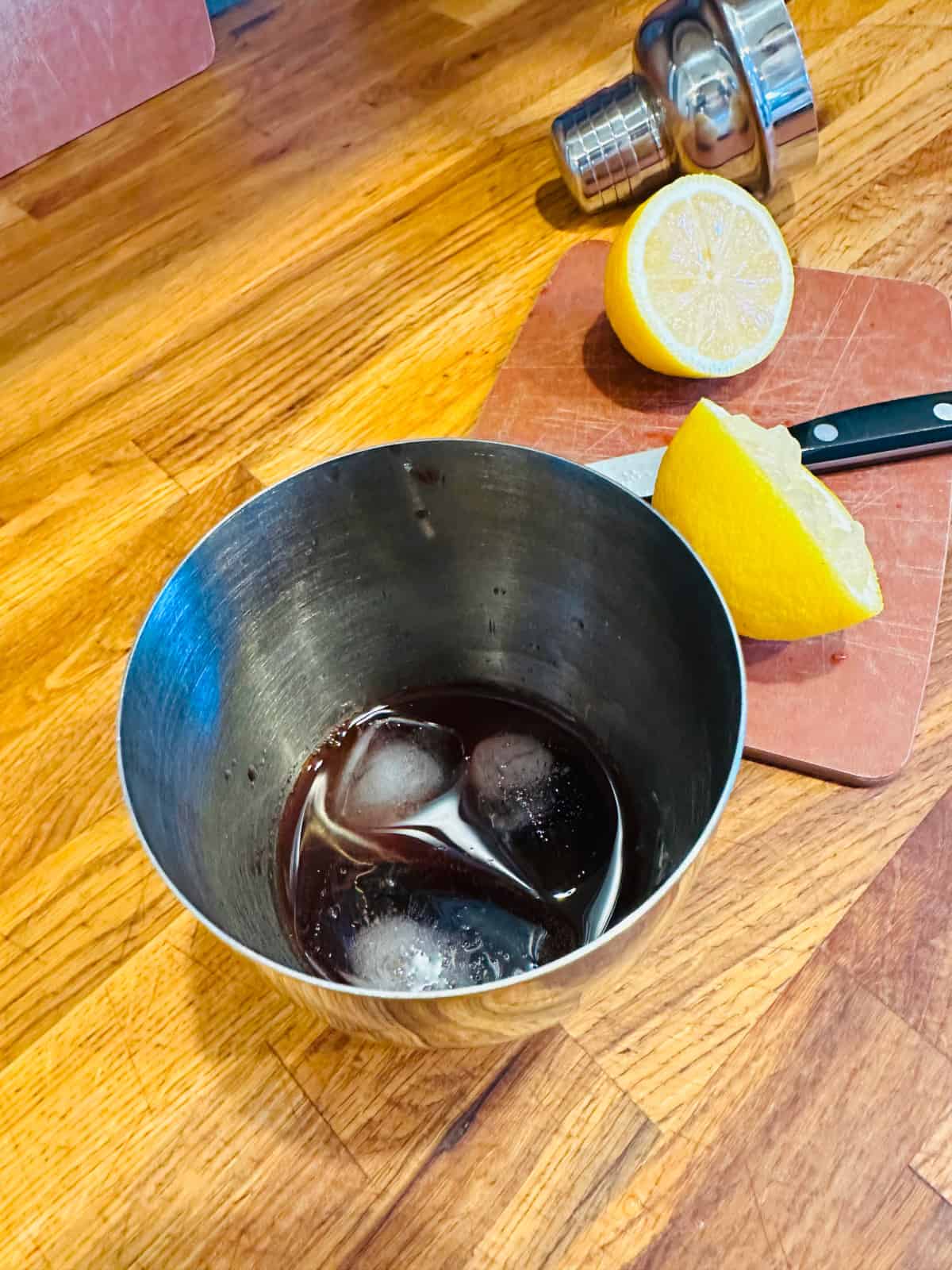 Metal cocktail shaker with ice and dark red liquid next to lemon cut in half.