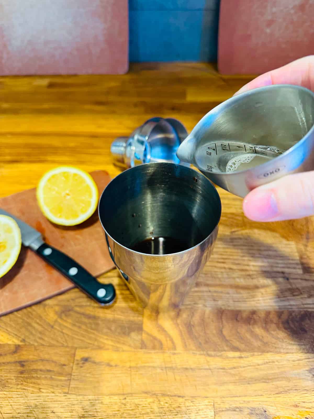 Hand pouring lemon juice from metal measuring cup into cocktail shaker.