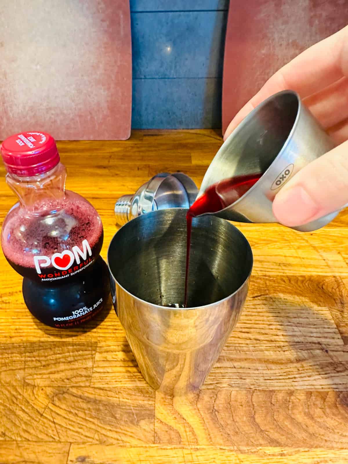 Hand pouring pomegranate juice from metal measuring cup into cocktail shaker.