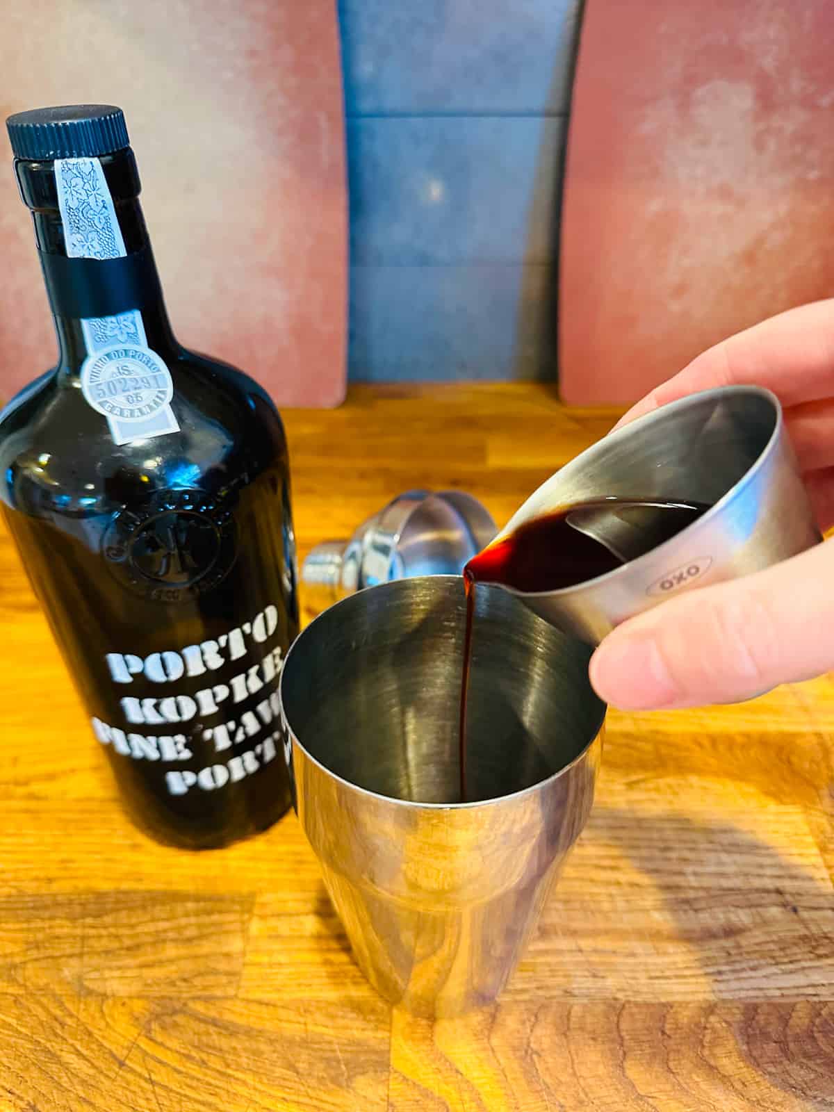 Hand pouring tawny port from metal measuring cup into cocktail shaker.