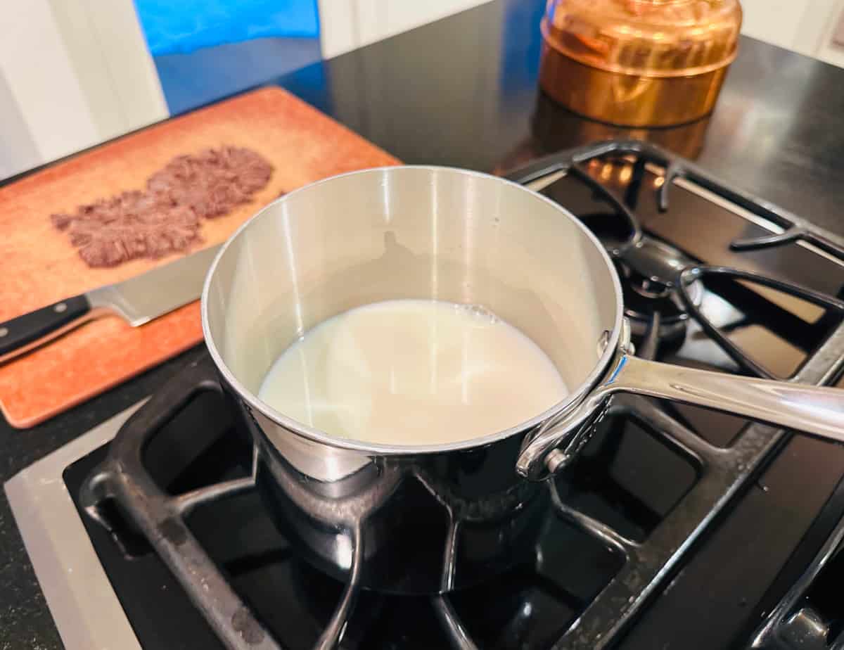 Milk in a small steel saucepan sitting on the stove next to chopped chocolate on a cutting board.