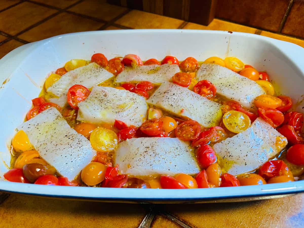 Raw fish fillets nestled into a bed of roasted tomatoes in a large white baking dish.