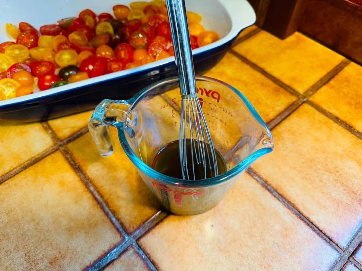 Olive oil and sherry vinegar mixture in a glass measuring cup with a metal whisk next to a baking dish full of halved tomatoes.