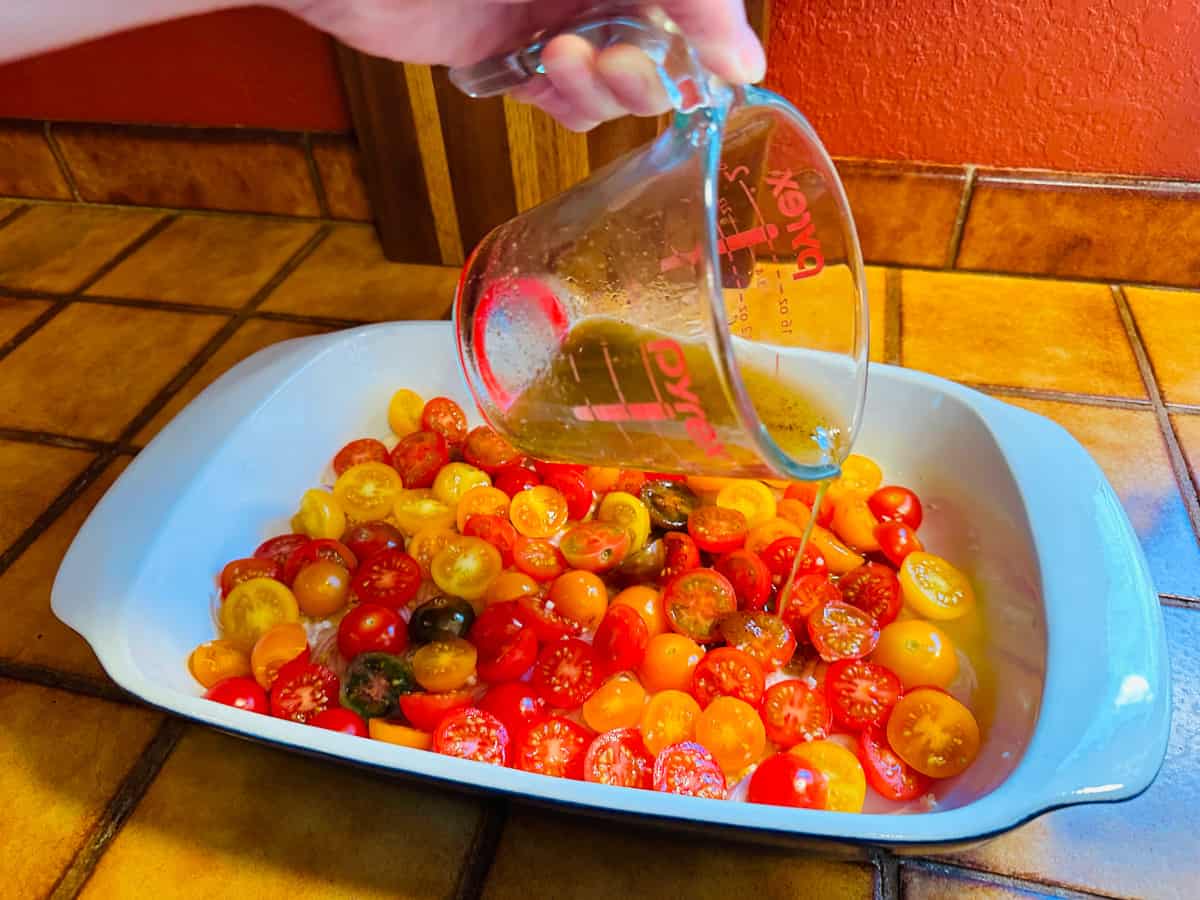 Oil and vinegar mixture being poured from a glass measuring cup into a baking dish full of halved cherry tomatoes.