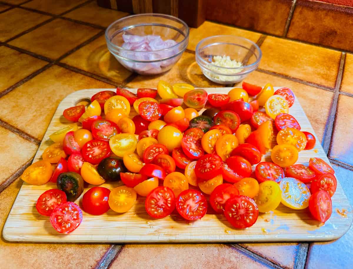 Halved cherry tomatoes covering a large cutting board and two small glass bowls containing chopped shallots and minced garlic.
