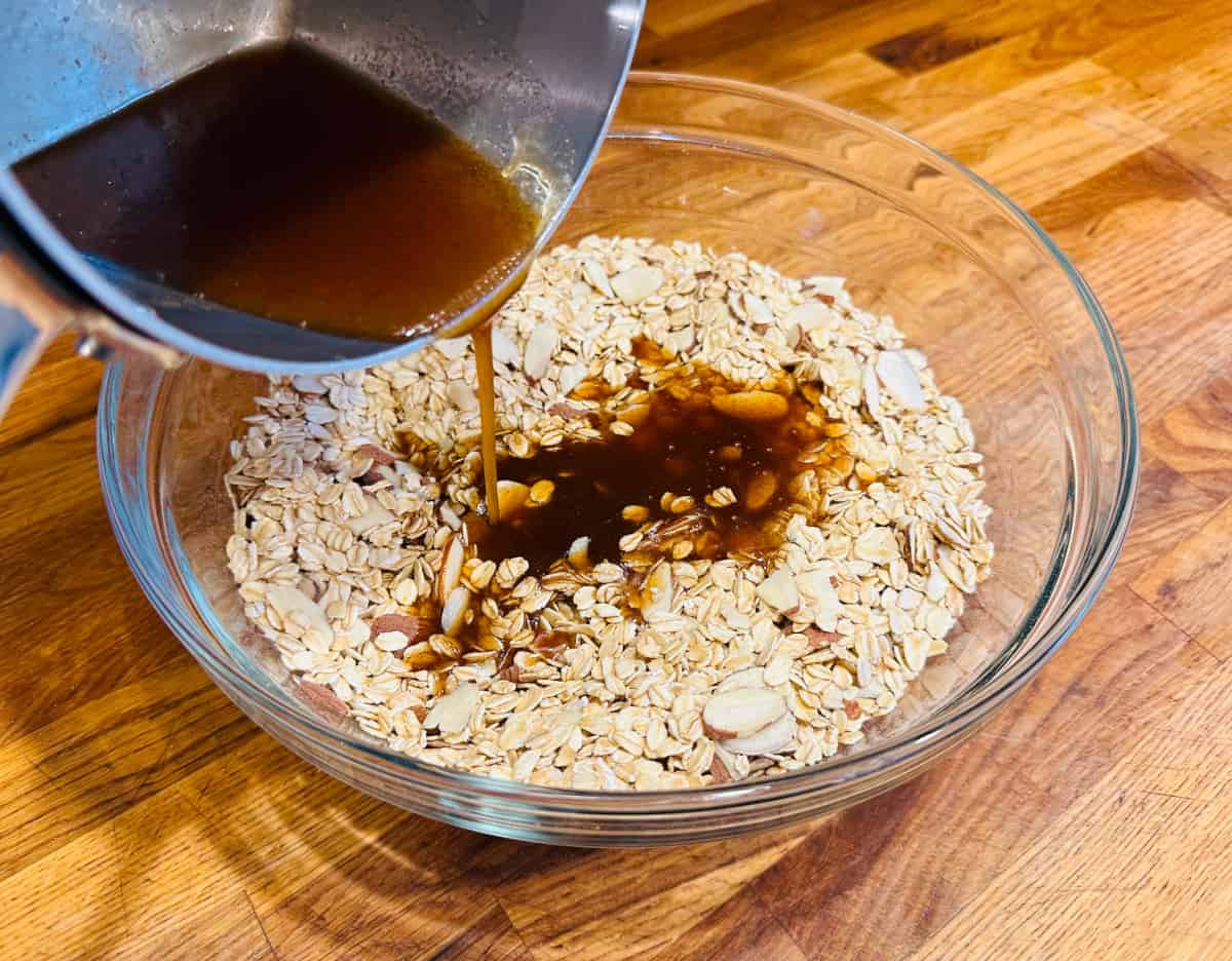 Pouring honey and butter mixture from metal saucepan into oats and almonds in a glass bowl.