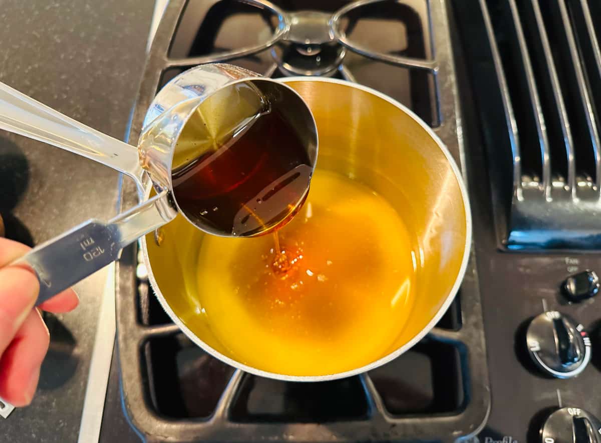 Pouring honey from a metal measuring cup into melted butter in a small metal saucepan.