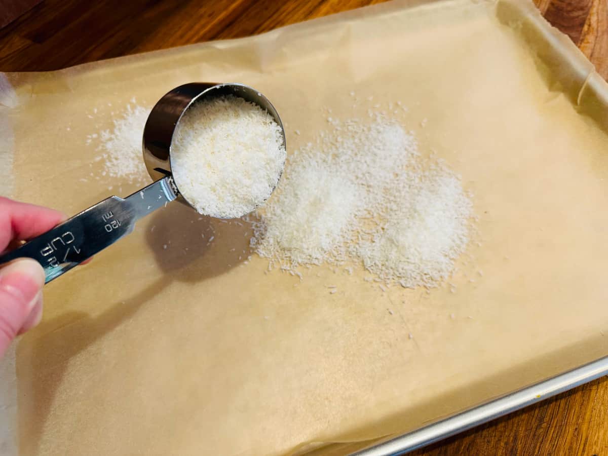 Spreading shredded coconut from a measuring cup onto a metal baking sheet covered with parchment paper.