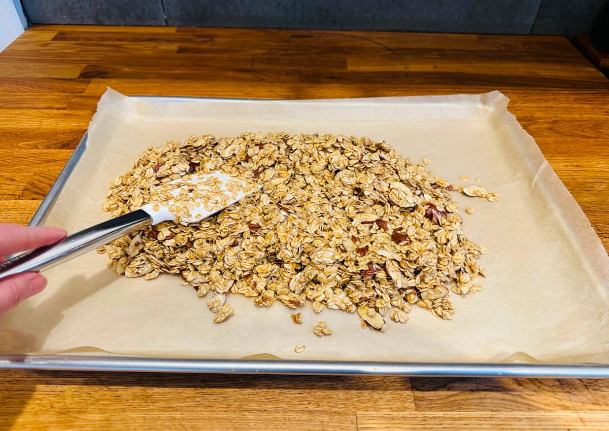 Spreading granola mixture with a white silicone spatula on a metal baking sheet covered with parchment paper.