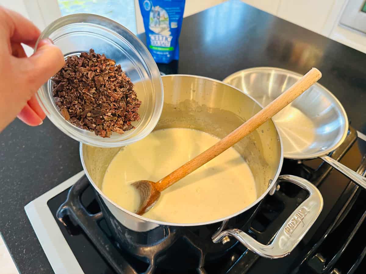 Adding cacao nibs to the milk mixture in a metal pot with a wooden spoon resting in it.