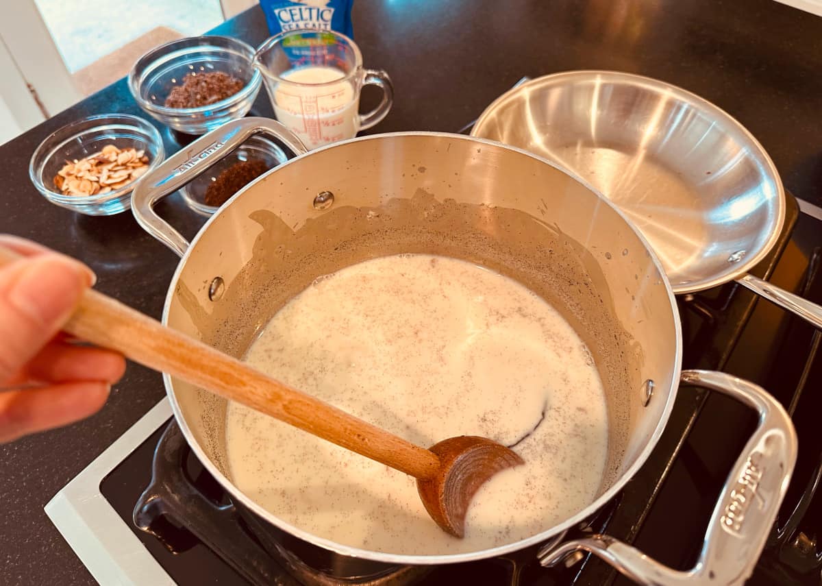 Milk mixture simmering in a metal pot while being stirred with a wooden spoon next to flavoring ingredients set out in small glass bowls on the counter.