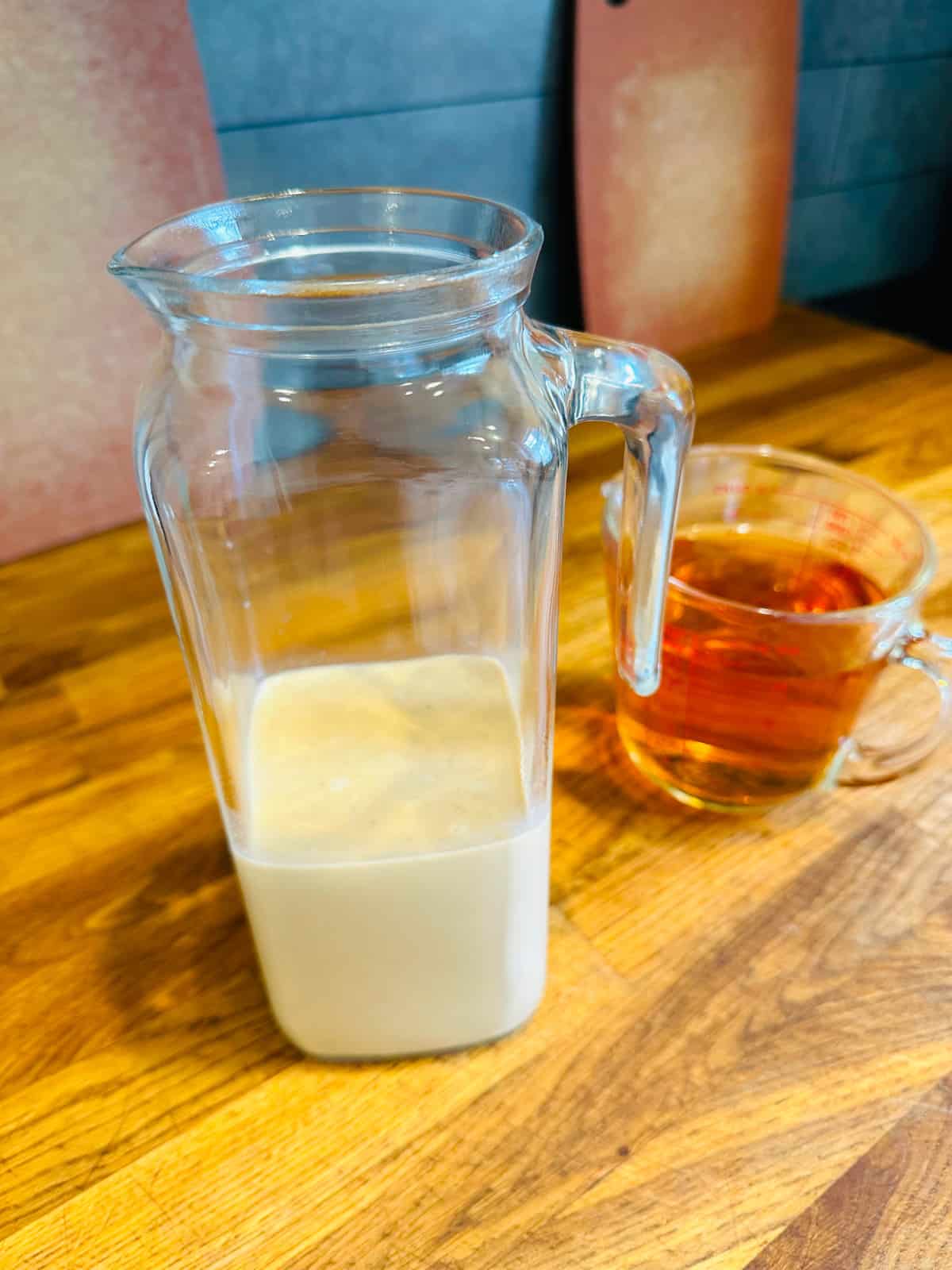 Tall glass pitcher filled half way up with pale creamy liquid next to a glass measuring cup filled with golden brown whiskey.