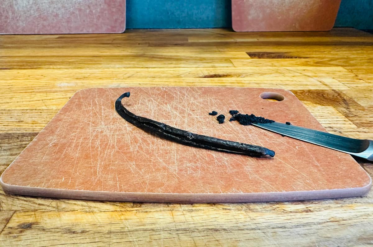 Split vanilla bean laying on a cutting board next to a small knife with vanilla seeds on the tip.