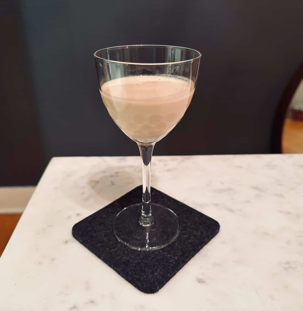 Irish cream in a small stemmed glass sitting on a dark gray coaster on a white marble table.