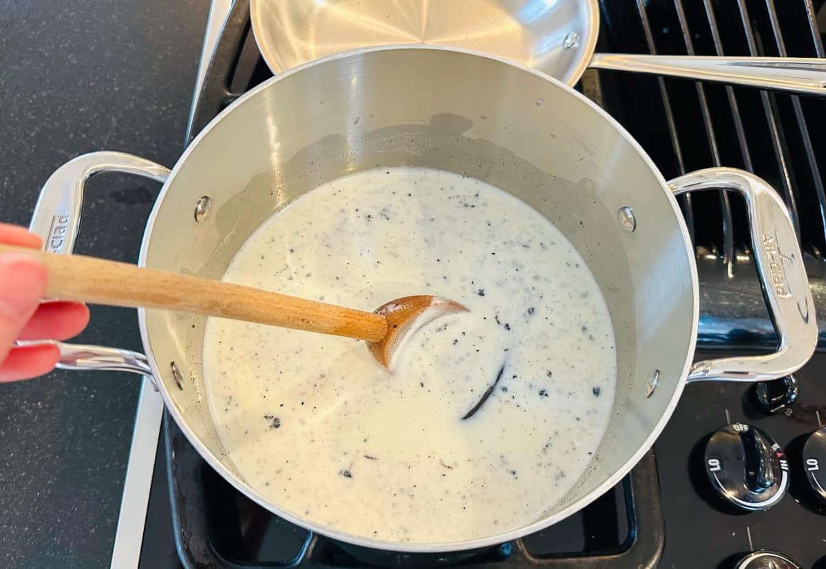 Milk, sugar, and vanilla bean simmering in a metal pot with a wooden spoon.