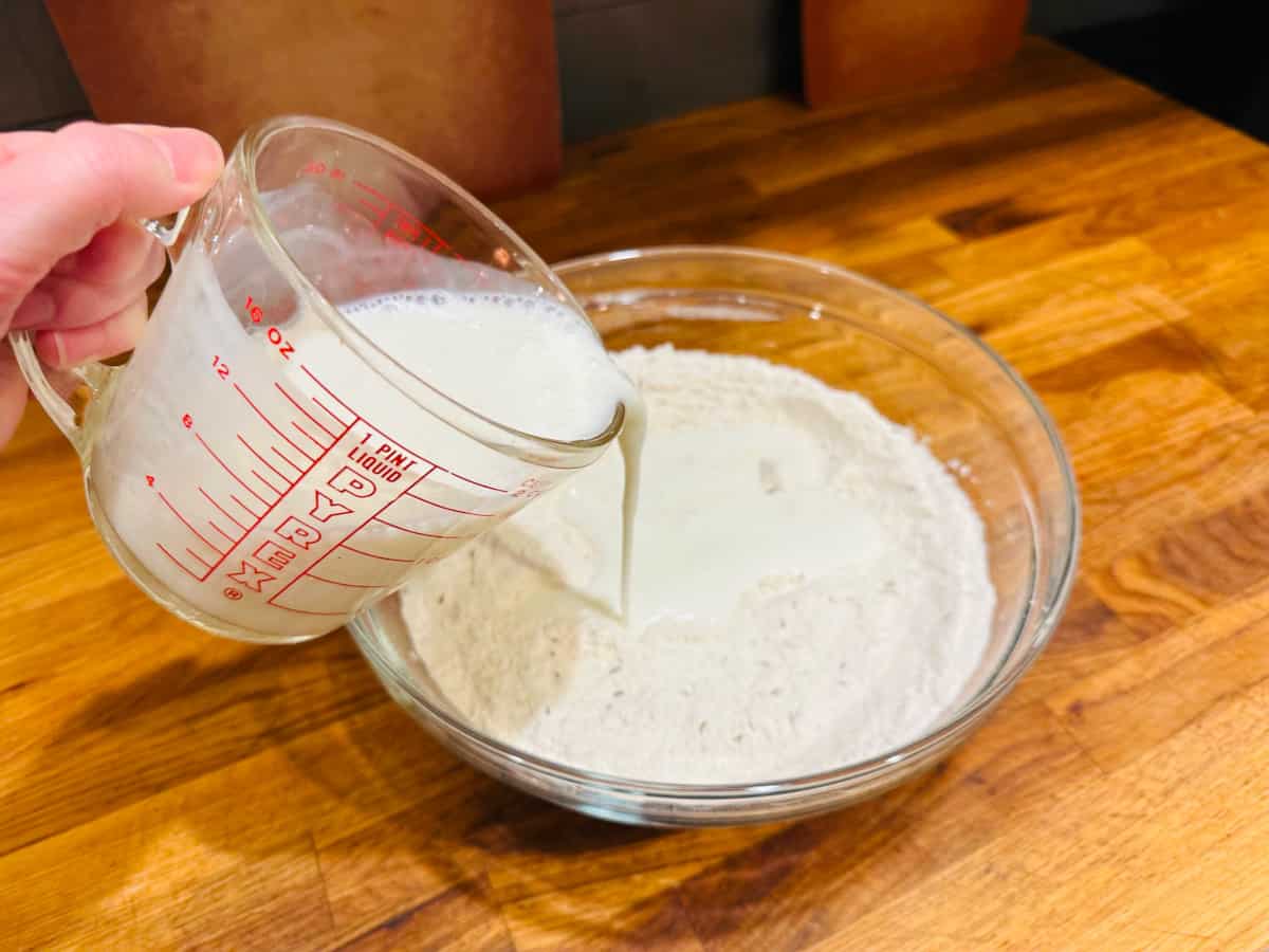Pouring buttermilk from a glass measuring cup into flour and dry ingredients in a glass bowl.